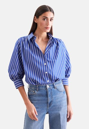 Vrouw met steil donker haar draagt een blauw-wit verticale gestreepte blouse met pofmouwen en een blauwe jeans met hoge taille.