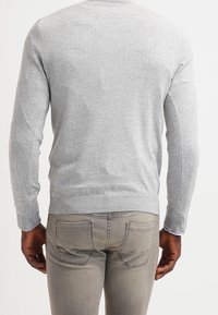 Man in een lichtgrijze trui en beige jeans, van achteren gezien tegen een simpele witte achtergrond.