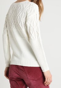 Femme portant un pull blanc en maille torsadée avec des motifs texturés sur les manches et le dos, associé à un pantalon en velours côtelé rouge foncé.