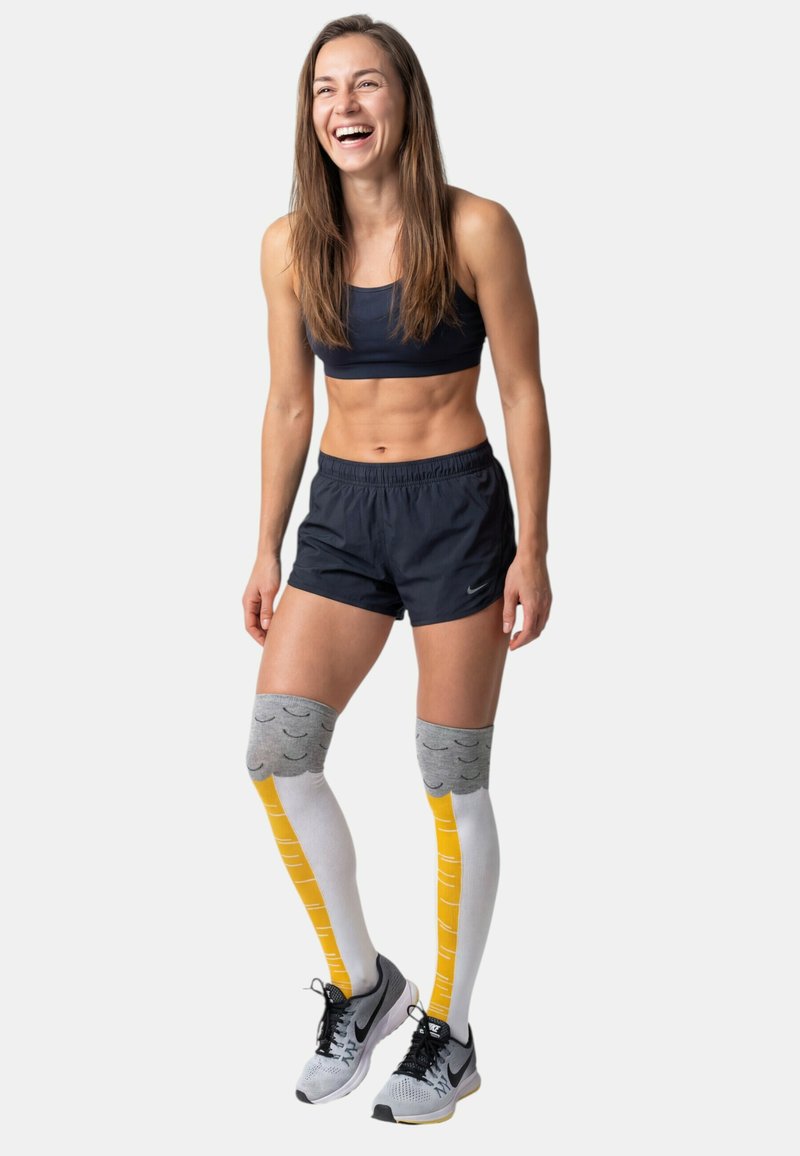 Femme athlétique en tenue de sport noire et chaussettes de compression montantes gris, blanc et jaune, souriante et debout devant un fond uni.