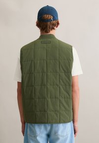 Zielona pikowana kamizelka bez rękawów, z tylną metką, noszona na białej koszuli, w połączeniu z jasnoniebieskimi dżinsami.