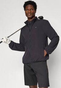 Giacca a vento nera con colletto alto e chiusura con zip. Indossata sopra un polo nera, abbinata a pantaloni shorts neri. Modello che tiene in mano un bastone da golf.