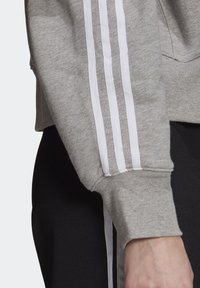 Grå sweatshirt med vita sidremsor. Tyget verkar mjukt med en ribbad mudd och synlig sömnad. Inga mönster. Accenter inkluderar dragsko i nederkant.