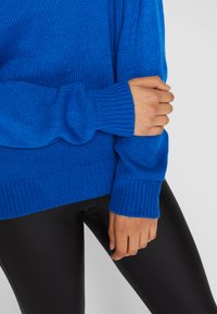 Strikket blå genser med ribbede kanter og mansjetter, som viser strukturen i stoffet; kombinert med svarte leggings.