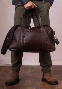 Brown leather duffel bag with dual handles, adjustable shoulder strap, and front zip pocket. Smooth texture, rectangular shape.