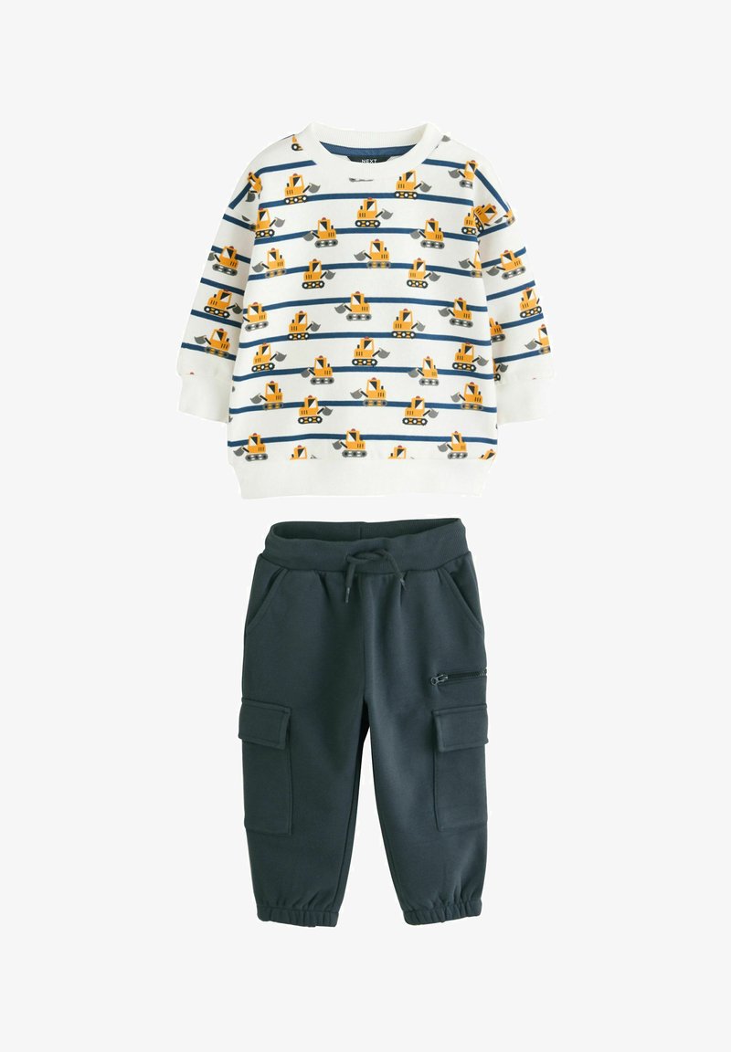 Biely sveter s modrými pruhmi a žltými vzormi bagerov, skombinovaný s tmavošedými cargo nohavicami s vreckami na zips a s nášivkami.