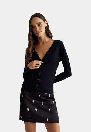 Cardigan nero con scollatura a V profonda, bordi in pizzo e chiusura a bottoni. Abbinato a una gonna nera con dettagli metallici sparsi.