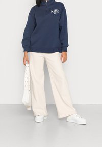 Marinblå Nike-tröja med hög krage och vit logo, kombinerad med vida krämfärgade byxor och vita plattformssneakers. Accessoarerna inkluderar en vit väska.