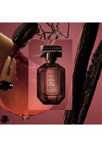 Mörk bärnstenfärgad glasparfymflaska med geometrisk design, ett gyllene lock och "BOSS"-branding, placerad mot en dämpad rosa bakgrund med frukt- och vätskeakenter.