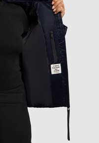 Námornícka puffer bunda zvnútra s hladkou textúrou, obsahujúca zipsovú kapsu a štítok s nápisom: "Teplé objatie z látky."