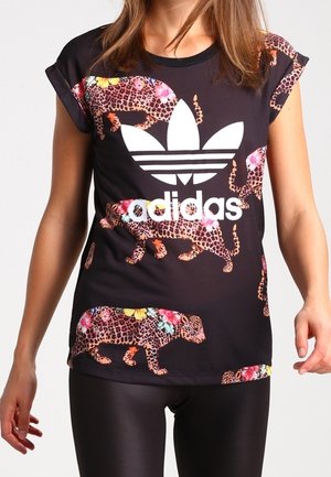 Femme portant un t-shirt Adidas noir avec un logo blanc et un imprimé léopard décoré de fleurs colorées, associé à des leggings noirs brillants.