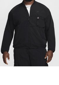 Man in een zwart Nike zip-up jack en bijpassende zwarte broek, geaccessoird met zilveren kettingen en een witte onderhuid zichtbaar.