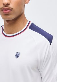 Νέος άνδρας που φοράει λευκό πουκάμισο K-Swiss με μπλε σκούρο πάνελ στους ώμους και ριγωτή λαιμόκοψη με κόκκινο, λευκό και μπλε χρώμα.