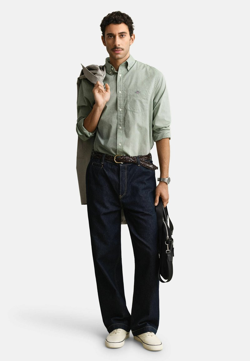 Hombre con camisa de botones verde claro, vaqueros oscuros, zapatos blancos, sosteniendo una chaqueta sobre el hombro y una bolsa negra en la mano.