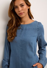 Džinsa kleita ar garām piedurknēm, apaļu kakla izgriezumu un brīvu piegriezumu. Zilā audumā ar nedaudz teksturētu virsmu un sānu kabatām.