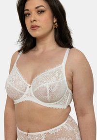 Soutien-gorge en dentelle blanche avec des bonnets souples, des bords festonnés et un détail de nœud central. Les bretelles réglables et la texture transparente rehaussent son design délicat.