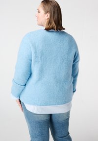 Lichtblauwe, zachte trui met lange mouwen, ronde hals en een losse pasvorm. Draagt over een wit met lichtblauw gestreept shirt en spijkerbroek.