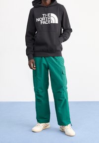 Una persona che indossa una felpa nera The North Face, pantaloni cargo color teal e sneaker crema, in piedi su un pavimento azzurro chiaro contro un muro bianco.