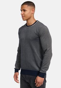 Strikkegenser i mørkegrått med subtil tekstur, rund hals, og mørkeblå ribbestrikkede mansjetter og nederkant. Har en kontrasterende lys trim.