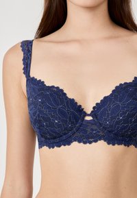 Navy blauwe kanten bh met golvende randen, dunne bandjes en een decoratieve uitsnijding in het midden. Bevat bloemmotieven en een lichte glans.