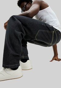 Jeans en denim foncé avec des détails de couture jaune, coupe droite, portés avec des baskets blanches. Le mannequin est accroupi, mettant en valeur le design du pantalon.