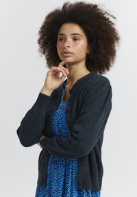 Mørkeblå strikket cardigan med en let struktur, rund halsudskæring og frontlukning med knapper. Båret over en blå mønstret kjole.