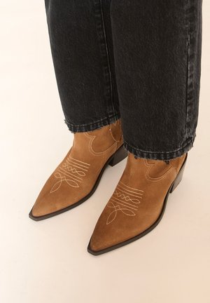 Bottines en daim marron à bout pointu avec des motifs décoratifs cousus, portées avec un jean noir à ourlet effiloché sur un sol clair.