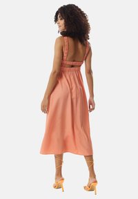 A mid-length, backless dress in soft coral fabric with ruffled straps and a gathered waist. Paired with orange strappy heels.