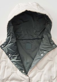 Beige Steppjacke mit einer dunkelgrünen gefütterten Kapuze, ausgestattet mit Druckknöpfen und einer strukturierten Oberfläche. Innenschild mit sichtbarem Logo.