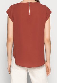 Rostfarbende Bluse mit kurzärmeligen, runden Ausschnitt und einem Reißverschluss im Rücken. Glattes Material mit seitlichen Schlitzen am Saum.
