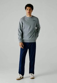 Jovem em pé, vestido com um moletom cinza da Red Bull Racing, jeans azul escuro e ténis brancos com solas de borracha.
