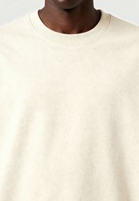 Šviesiai smėlinė purus džemperis su apvalia iškirpte, pagamintas iš švelnaus, tekstūrinio audinio. Sukurtas su raukšlėta iškirpte ir šiek tiek taškų visame paviršiuje.