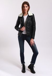 Black leather motorcycle jacket with a white shearling collar, paired with a white shirt and dark blue jeans. Model stands in a neutral background.