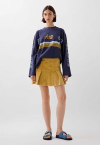 Sudadera larga de manga morada con rayas amarillas y azules; falda mini de pana amarilla con pliegues; sandalias azules con detalles decorativos.
