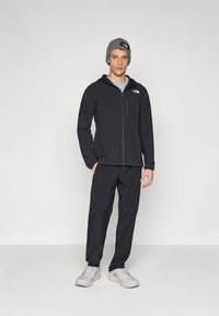 Schwarze Jacke mit Reißverschluss und Logo, kombiniert mit passenden Hosen und hellgrauen Schuhen. Das Model trägt eine graue Mütze. Minimalistisches Design.
