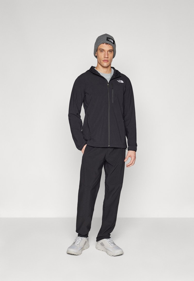 Schwarze Jacke mit Reißverschluss und Logo, kombiniert mit passenden Hosen und hellgrauen Schuhen. Das Model trägt eine graue Mütze. Minimalistisches Design.