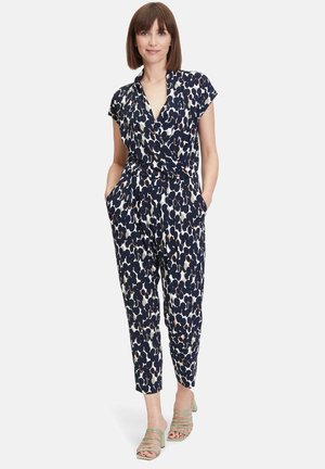 Tuta navy con stampa astratta in crema e marrone, maniche corte, vita cintrata e tasche, abbinata a sandali verdi con cinturini.