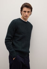 Dunkelgrüner, kabelgestrickter Pullover mit rundem Ausschnitt sowie gerippten Bündchen und Saum. Getragen über dunkelblauen Hosen vor neutralem Hintergrund.