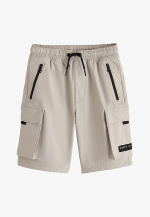 Person iført beige cargoshorts, hvide sokker og hvide sneakers, der står mod en neutral baggrund, set bagfra.