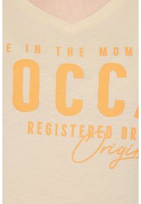 Camiseta de cuello en V beige con gran texto naranja que dice "OCC" y "Marca Registrada Original," con una textura suave y un estilo casual.