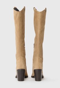 Bottes en suède beige jusqu'au genou avec une forme effilée, zips latéraux et talons en bois empilés. Présente une texture lisse et des coutures distinctes.