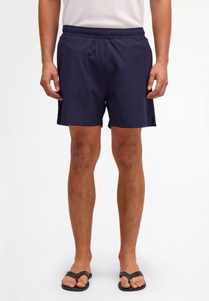 Mand iført marineblå shorts med elastik i taljen, hvid t-shirt og sorte klipklappere, stående mod en ensfarvet lys baggrund.