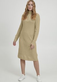 Beige gebreide jurk met een hoge hals en lange mouwen, voorzien van een textuurpatroon en zijsplitten, gecombineerd met witte sneakers.
