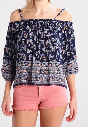 Vrouw draagt een marineblauw off-shoulder blouse met bloemenprint en driekwart mouwen, gecombineerd met roze denim shorts met rafelige randen.