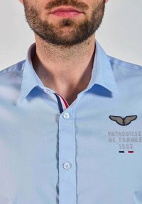 Chemise boutonnée bleu clair avec un col pointu, comportant un logo et du texte brodés, ainsi qu'un détail de col rayé en rouge, blanc et bleu.