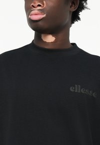 Sort sweatshirt med rund halsudskæring og et tonalt logo "ellesse" på venstre side. Materialet virker blødt med en glat tekstur.