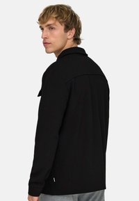 Junger Mann mit blondem Haar, der ein schwarzes Langarmshirt trägt, leicht zur Seite gedreht, vor einem schlichten hellen Hintergrund.