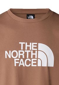 Camiseta de algodón marrón con cuello redondo, que presenta un gran logo blanco con formas curvas. Textura suave, diseño casual, detalles mínimos.