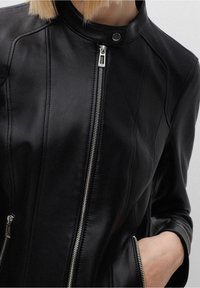 Czarna skórzana kurtka z srebrnym zamkiem błyskawicznym i napami przy kołnierzu, noszona przez osobę z ręką w zapiętej kieszeni, przycięta poniżej podbródka i w talii.