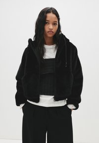 Črna umetna krzna jakna s kapuco, z zadrgo in stranskimi žepi. Oblečena čez slojevito črno-belo obleko z rebrasto teksturo.